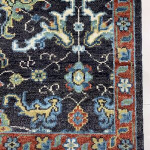 Navy blue rug corner showing colorful traditional motifs, a thick red border, and white fringe on white flooring.