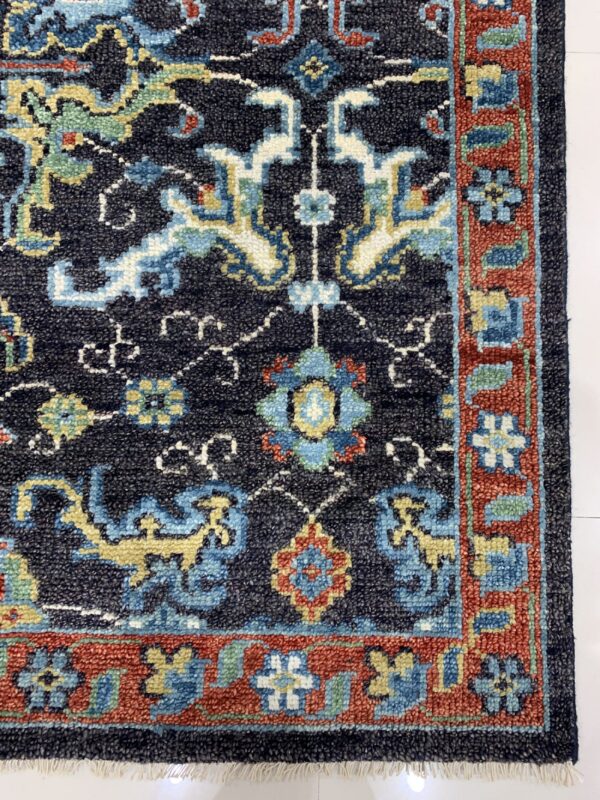 Navy blue rug corner showing colorful traditional motifs, a thick red border, and white fringe on white flooring.