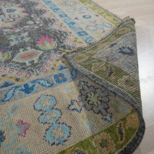 A multicolored traditional rug in gray, blue, and pink is rolled up, showing its textured backing on light wood floors.
