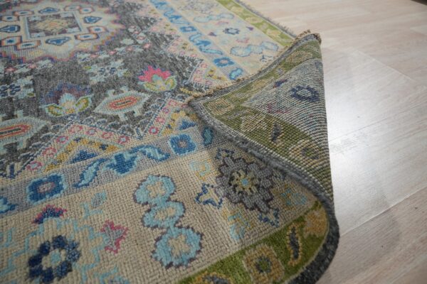 A multicolored traditional rug in gray, blue, and pink is rolled up, showing its textured backing on light wood floors.