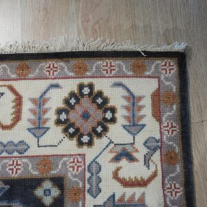 Geometric and stylized rug with blue, gray, and orange motifs on a cream field and light wood floor.