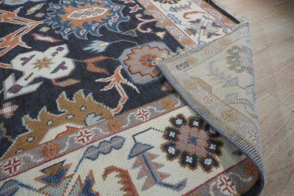 The edge of a dark patterned rug with cream and gray motifs is folded back on light wood flooring.