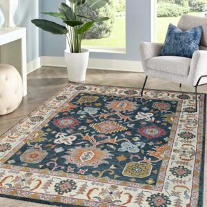 Traditional blue, cream, and orange rug on herringbone floor beside a gray chair and large potted plant.