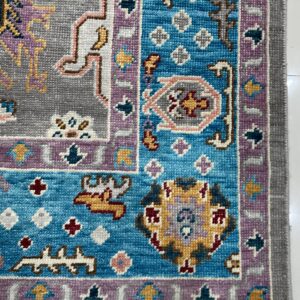 Intricate blue, gray, and purple geometric patterned rug corner with fringe displayed on white tile.