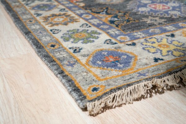 Corner view of a traditional gray, blue, and gold rug border with fringes on light wood floor.