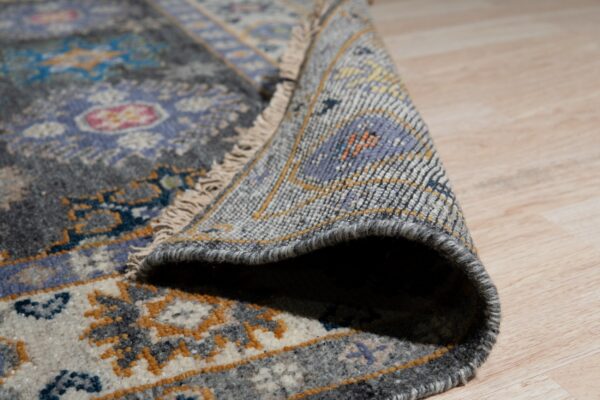 Close-up shows a rolled gray and lavender patterned rug with fringe on light wood flooring.