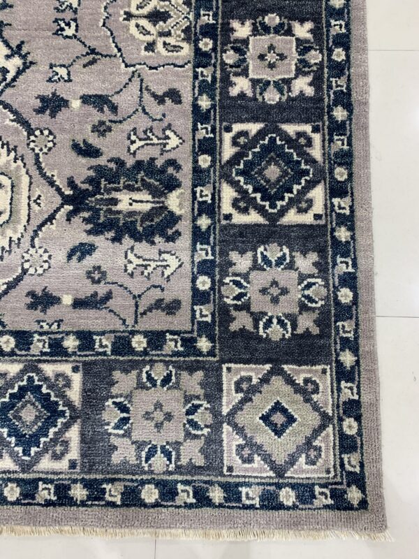 Close view of a textured lavender and navy rug border with geometric and floral patterns and white fringe on a tile floor.
