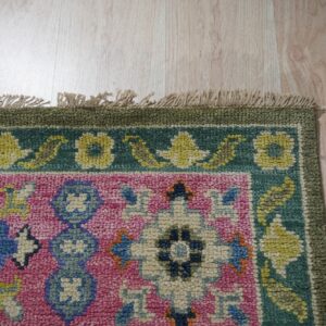 Colorful pink, blue, and green textured rug with floral patterns and visible fringe rests on light wood flooring.