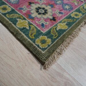 Bright pink rug corner featuring floral patterns, bordered by teal and olive green, sitting on light wood floor.