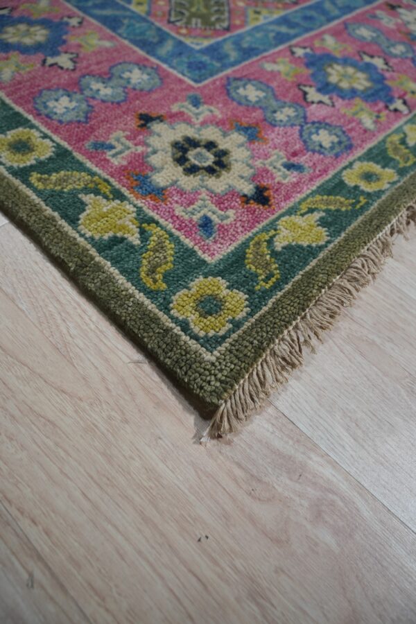 Bright pink rug corner featuring floral patterns, bordered by teal and olive green, sitting on light wood floor.