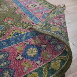 Curled traditional rug with olive green, bright pink, and blue geometric patterns on light wood floors.