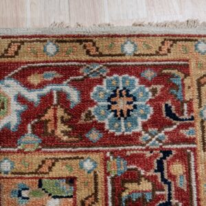 Textured rug corner displaying traditional red, gold, and blue floral patterns beside pale wood plank flooring.
