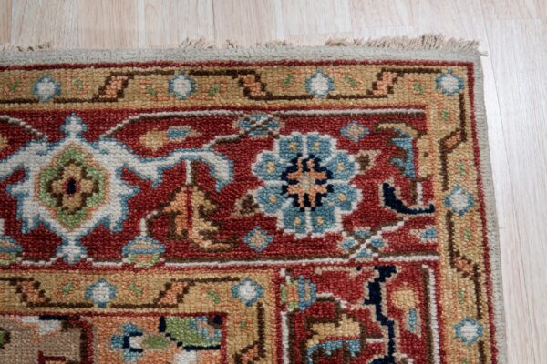 Textured rug corner displaying traditional red, gold, and blue floral patterns beside pale wood plank flooring.