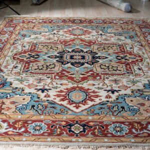 Cream, rust, and blue traditional rug with fringe and an intricate geometric design displayed on light wood flooring.