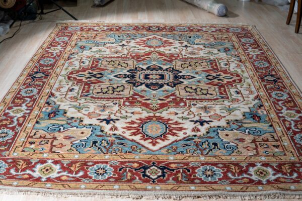 Cream, rust, and blue traditional rug with fringe and an intricate geometric design displayed on light wood flooring.