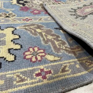 Close-up of a blue textured rug with yellow, gray, and maroon traditional patterns, showing the woven backing on wood flooring.