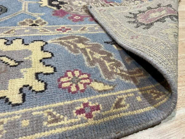 Close-up of a blue textured rug with yellow, gray, and maroon traditional patterns, showing the woven backing on wood flooring.