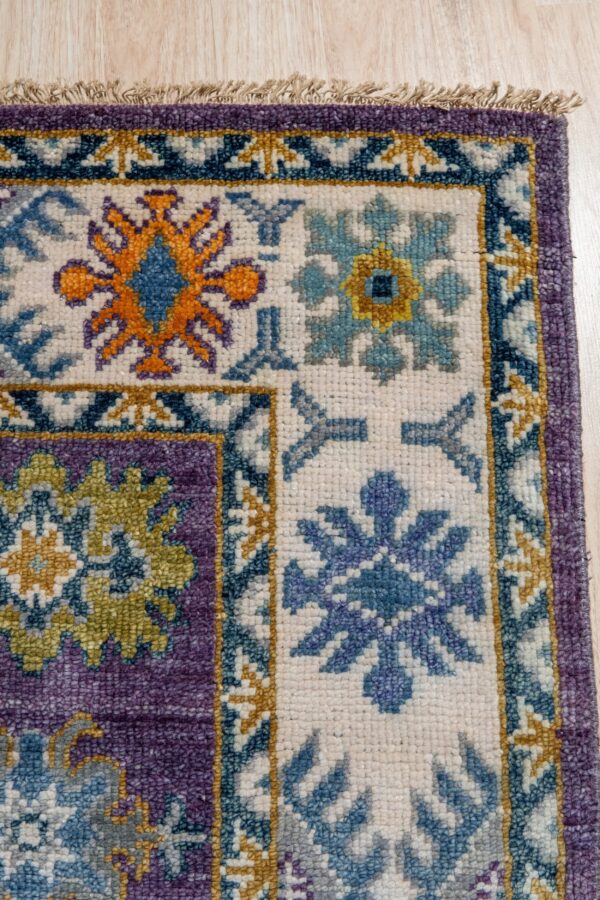 Geometric short-pile rug corner in purple, cream, teal, and orange resting on light wood flooring.