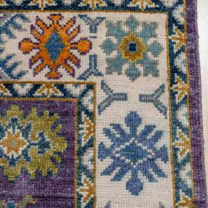 Detail shot of a rug corner showing purple, cream, orange, and blue motifs on light wooden floor.