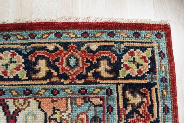 Red and navy high-pile rug with a detailed geometric border rests on light wood flooring.