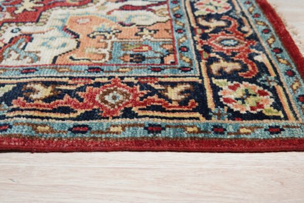 Traditional rug corner displaying red, navy, peach, and teal geometric patterns on light wood flooring.