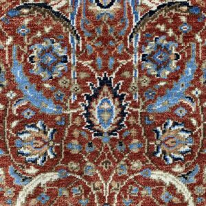 Textured close-up of a traditional red rug featuring intricate blue, cream, and brown ornamental motifs.