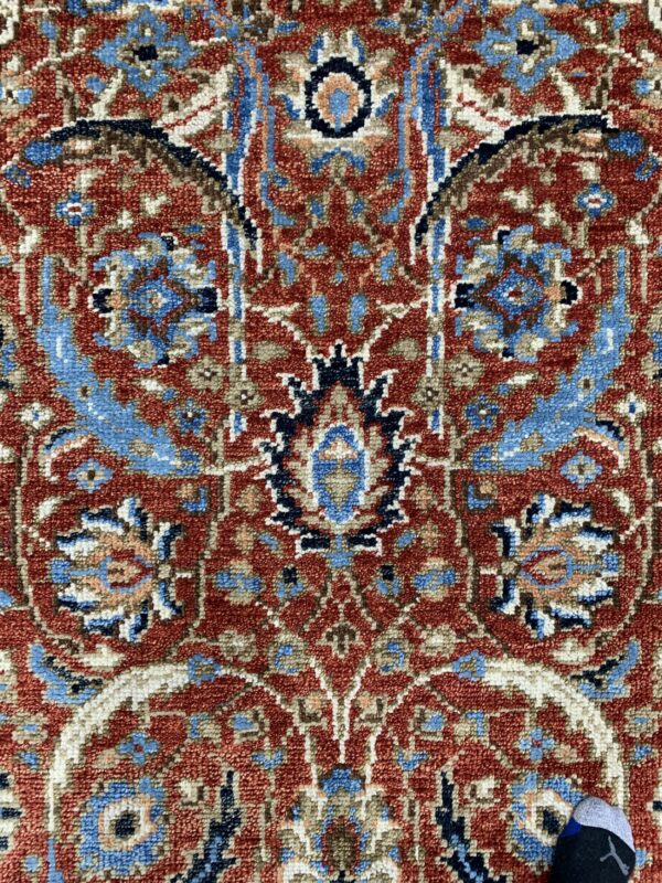 Textured close-up of a traditional red rug featuring intricate blue, cream, and brown ornamental motifs.