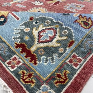 Corner of a blue, red, and grey patterned rug folded over a dusty rose patterned rug on white flooring.