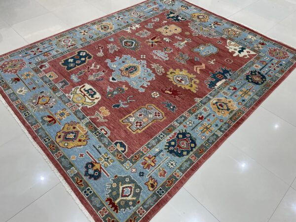 A rust-red rug with a low pile and colorful abstract folk motifs, featuring a wide light blue border, rests on white tile flooring.