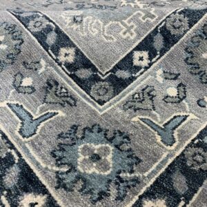 Textured high-pile rug in shades of grey, navy, teal, and cream displays complex traditional floral and geometric patterns.
