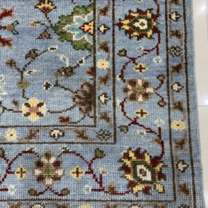 Textured light blue rug with a traditional multi-color floral pattern and dark brown border on white tile.