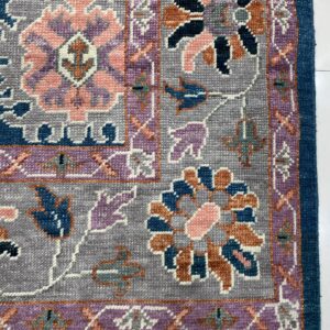 Detailed view of a rug corner with blue, gray, pink, and purple floral motifs and white fringe on tile.