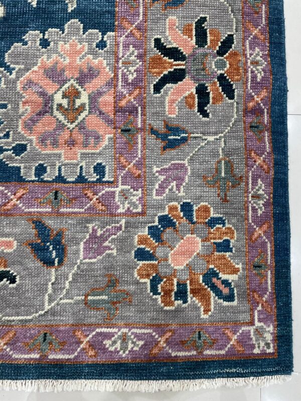 Detailed view of a rug corner with blue, gray, pink, and purple floral motifs and white fringe on tile.
