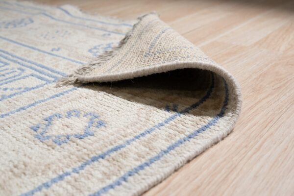 The rolled edge of a cream shag rug with blue geometric lines sits on light wood flooring.