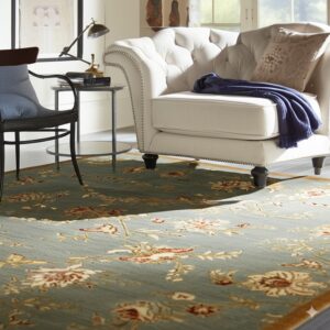 Blue-green floral rug with cream and rust accents beneath a white tufted chair and glass side table.