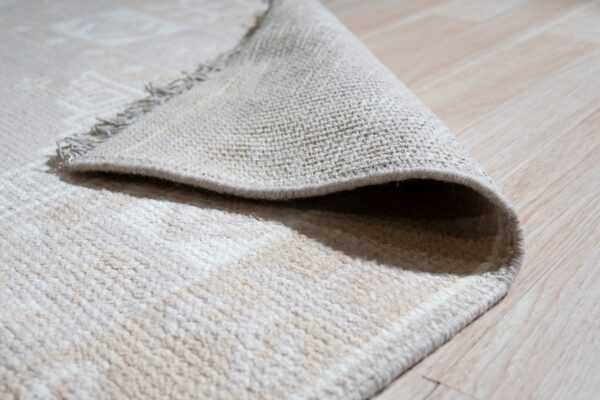 A cream, plush pile rug is folded over, showing its tightly woven backing on light wood flooring.