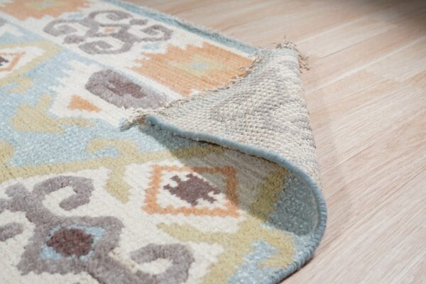 A medium-pile rug with abstract blue, peach, and taupe patterns is shown curled up on light wood flooring.