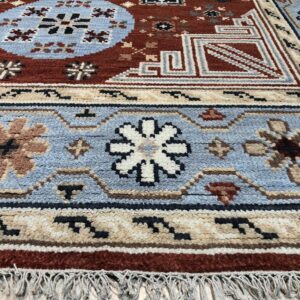 Textured rug in rust, blue, and tan with intricate geometric and floral patterns and visible fringe on light wood flooring.