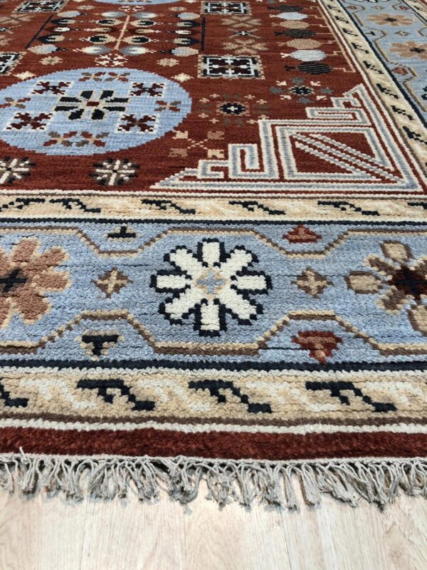 Textured rug in rust, blue, and tan with intricate geometric and floral patterns and visible fringe on light wood flooring.