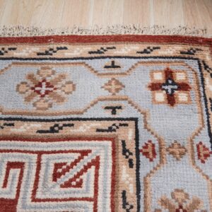 Corner detail of a blue, red, and beige geometric border rug with fringes on light wood planks.