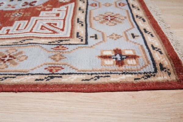 Rust, light blue, and beige geometric rug detail with fringe lying on light wood plank flooring.