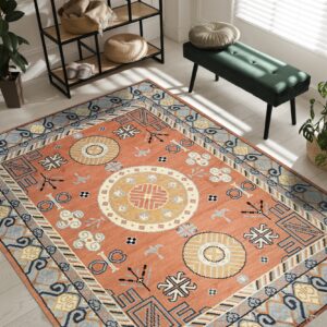Burnt orange rug with blue and yellow geometric patterns on light wood flooring next to a dark green bench.