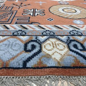 Textured area rug with rust field and blue tribal borders resting on light wood flooring.