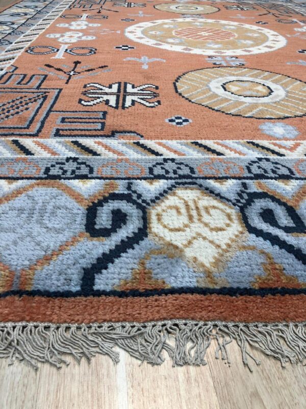 Textured area rug with rust field and blue tribal borders resting on light wood flooring.