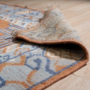 Rust and blue patterned rug is partially rolled, showing its woven backing on light wood flooring.