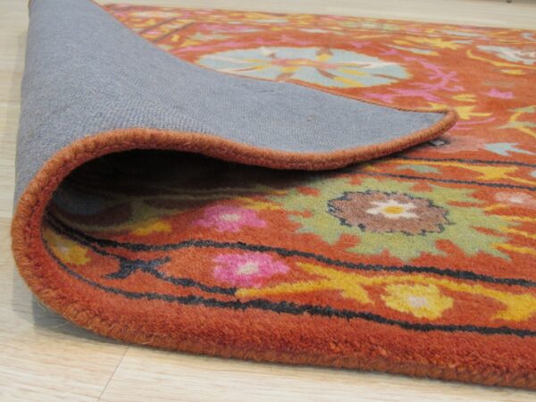 Rolled back rug corner reveals the gray backing and thick pile with orange, pink, and green floral patterns.