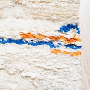 High-pile cream rug with thick shaggy texture and horizontal stripes of bright blue and orange on light wood flooring.