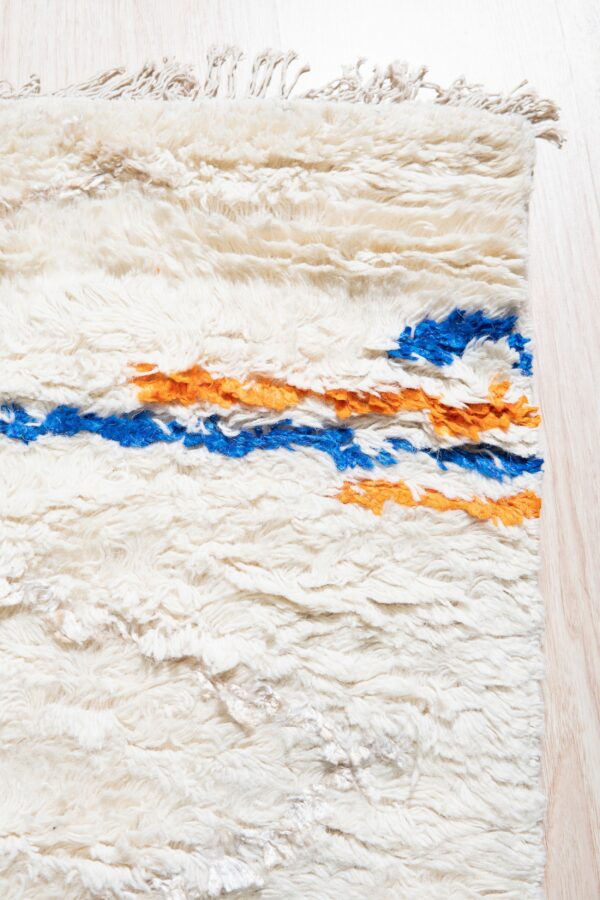 High-pile cream rug with thick shaggy texture and horizontal stripes of bright blue and orange on light wood flooring.