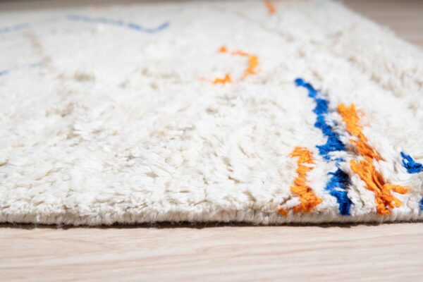 A shaggy cream rug with high pile displays abstract blue and orange patterns on light wood flooring.