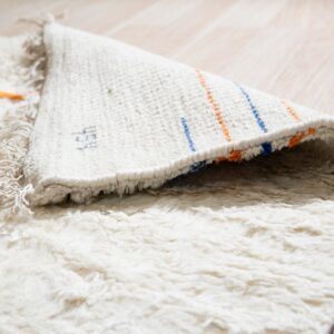 High-pile cream rug with orange accents folded over, showing a woven backing with blue and orange stripes on light wood flooring.